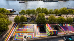 Aerial view of a colourful geometric rooftop sculpture garden beside the River Thames, surrounded by trees and boats in London.