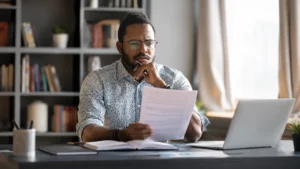 Man reviewing financial documents at a desk while checking savings and benefit eligibility information on a laptop