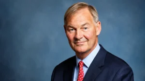 Professional wide corporate portrait of a senior businessman in a navy suit, light blue shirt and red patterned tie against a soft blue studio background, looking confidently at the camera