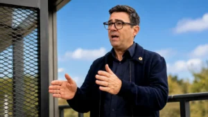 Wide editorial image of a man speaking outdoors with expressive hand gestures beside a modern metal structure under a clear blue sky.