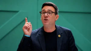 Wide editorial image of a man speaking with his index finger raised against a modern teal geometric background during a public address or conference