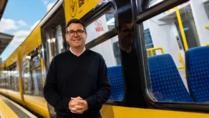 Wide editorial image of a smiling man standing beside a modern yellow passenger train at a railway station platform, with the train interior visible through the windows