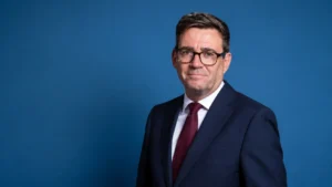 Wide professional portrait of a businessman in a navy suit and burgundy tie standing against a modern blue studio background with ample copy space