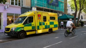 Wide street scene showing an emergency ambulance parked outside city buildings as a cyclist passes by on a sunny urban road.