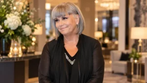 Silver-haired woman in a black outfit smiling in a warmly lit luxury lobby with flowers and contemporary furnishings.